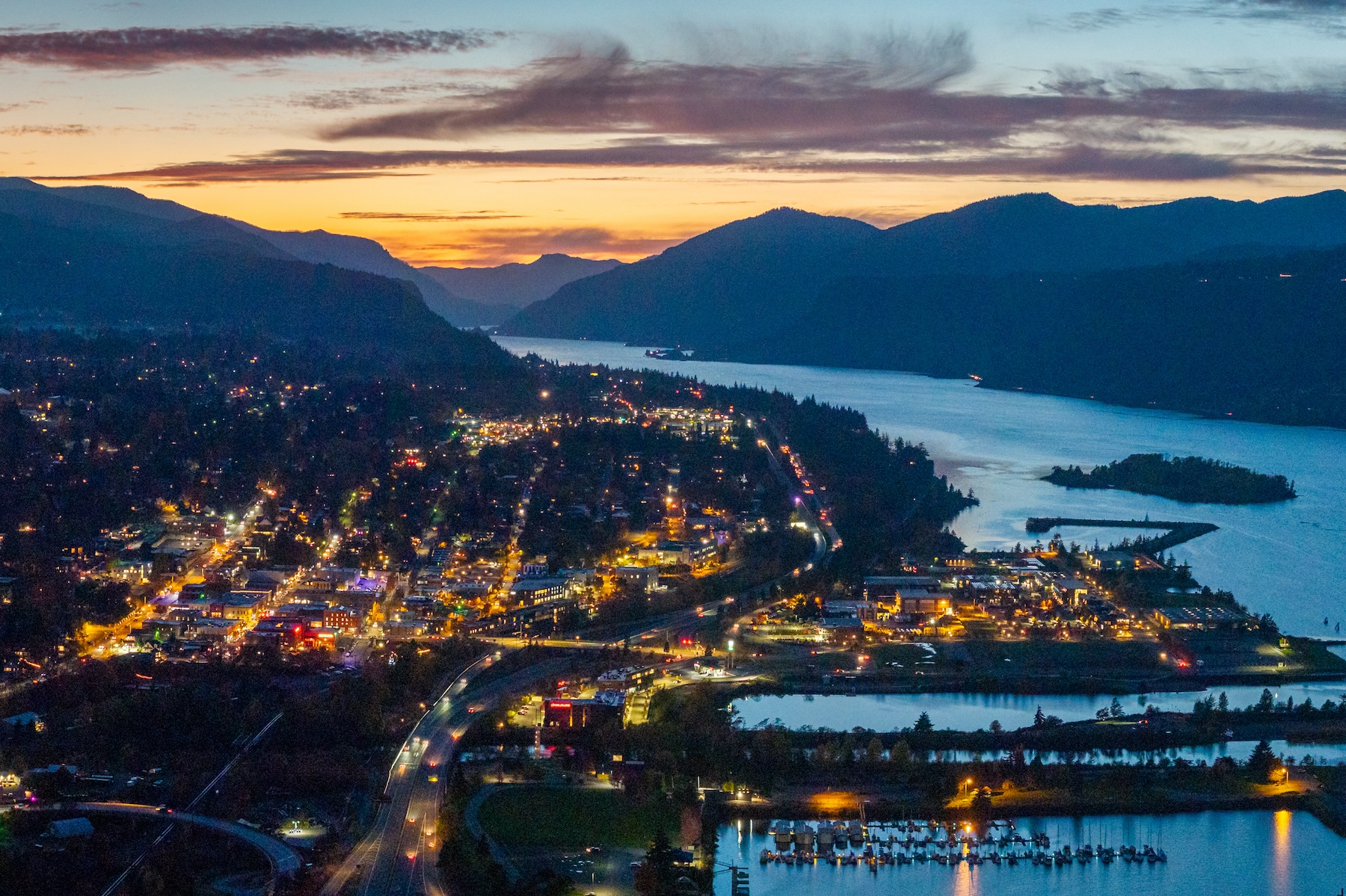 Aerial view of Hood River at sunset with city lights, the Columbia River, and surrounding mountains, showcasing the scenic location near Lightwell Hotel.