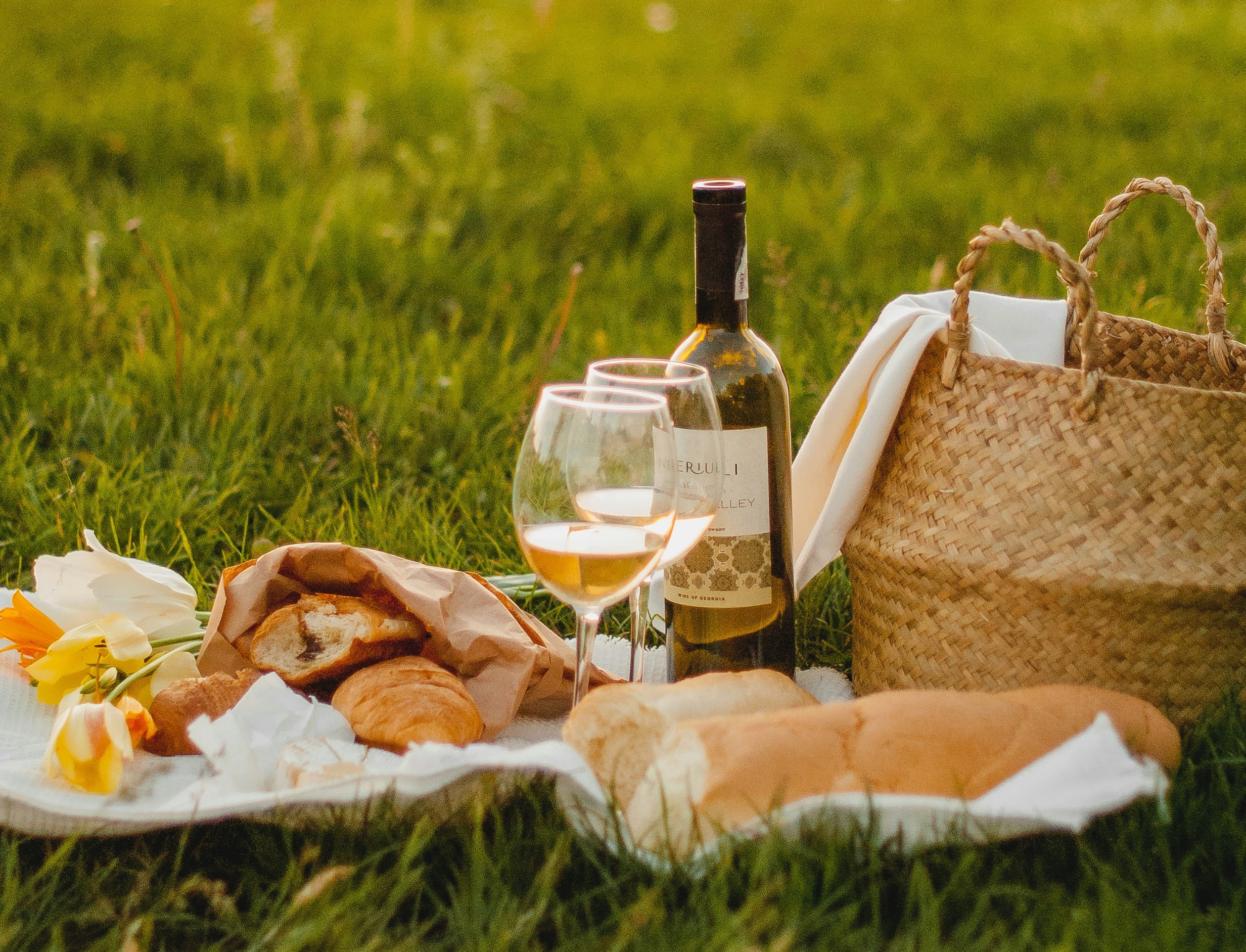 Picnic setup with white wine, bread, croissants, and flowers on a grassy field with a woven basket.