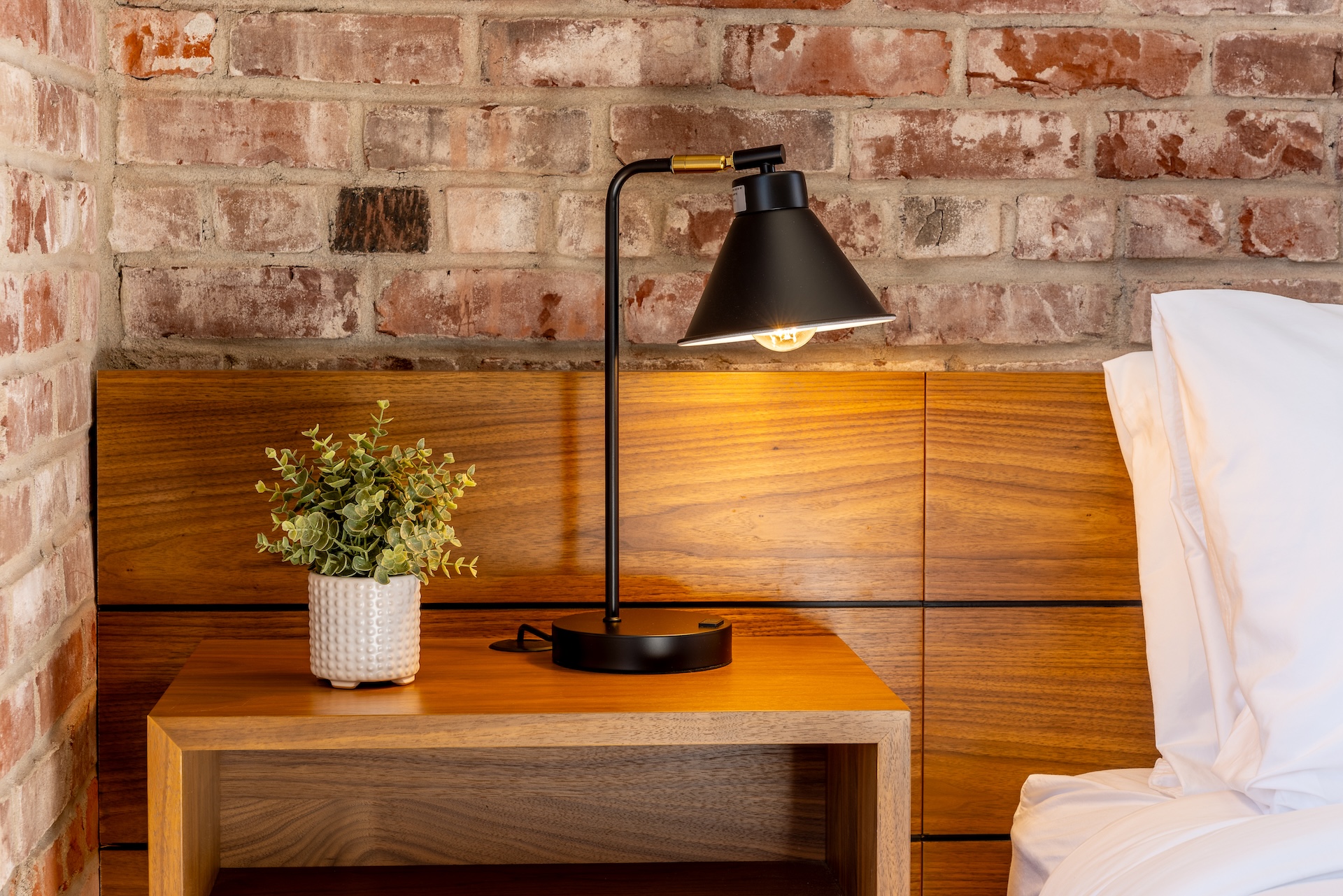 Lightwell Hotel & Spa guest room detail with black reading lamp, wooden nightstand, potted plant, and exposed brick wall in Hood River.