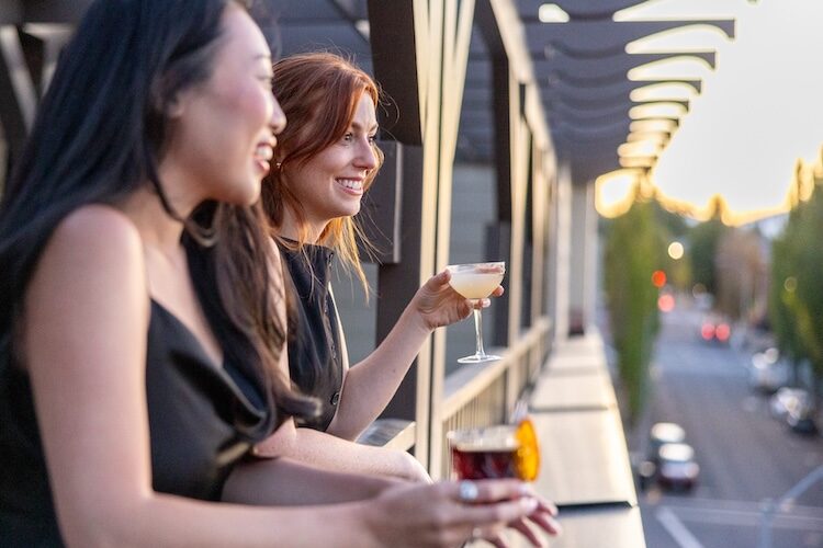 Two women enjoying cocktails at The Rooftop at Lightwell Hotel in downtown Hood River with sunset city views.